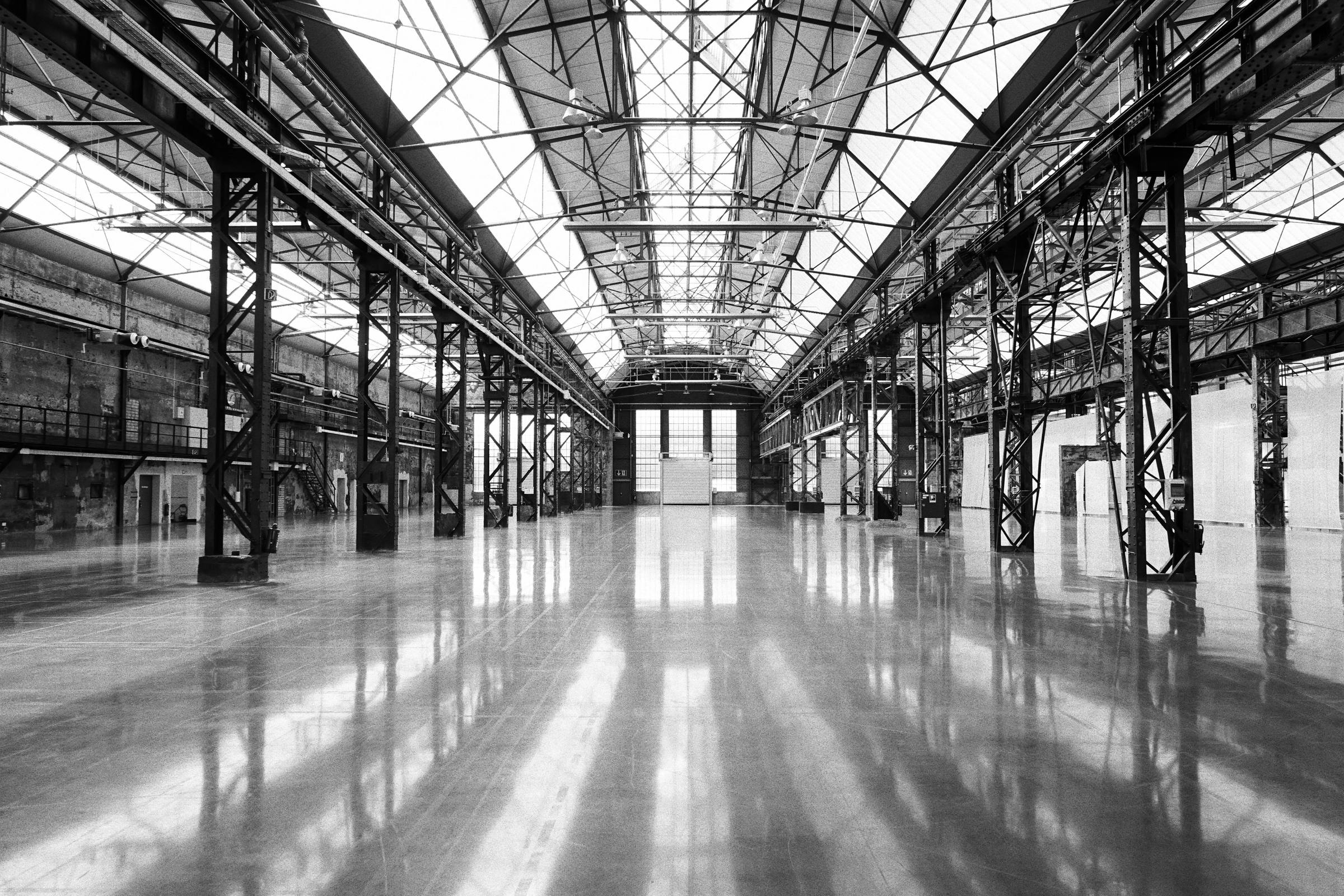 Black-and-white photograph of a long, empty steel hall with a glass roof and a polished concrete floor